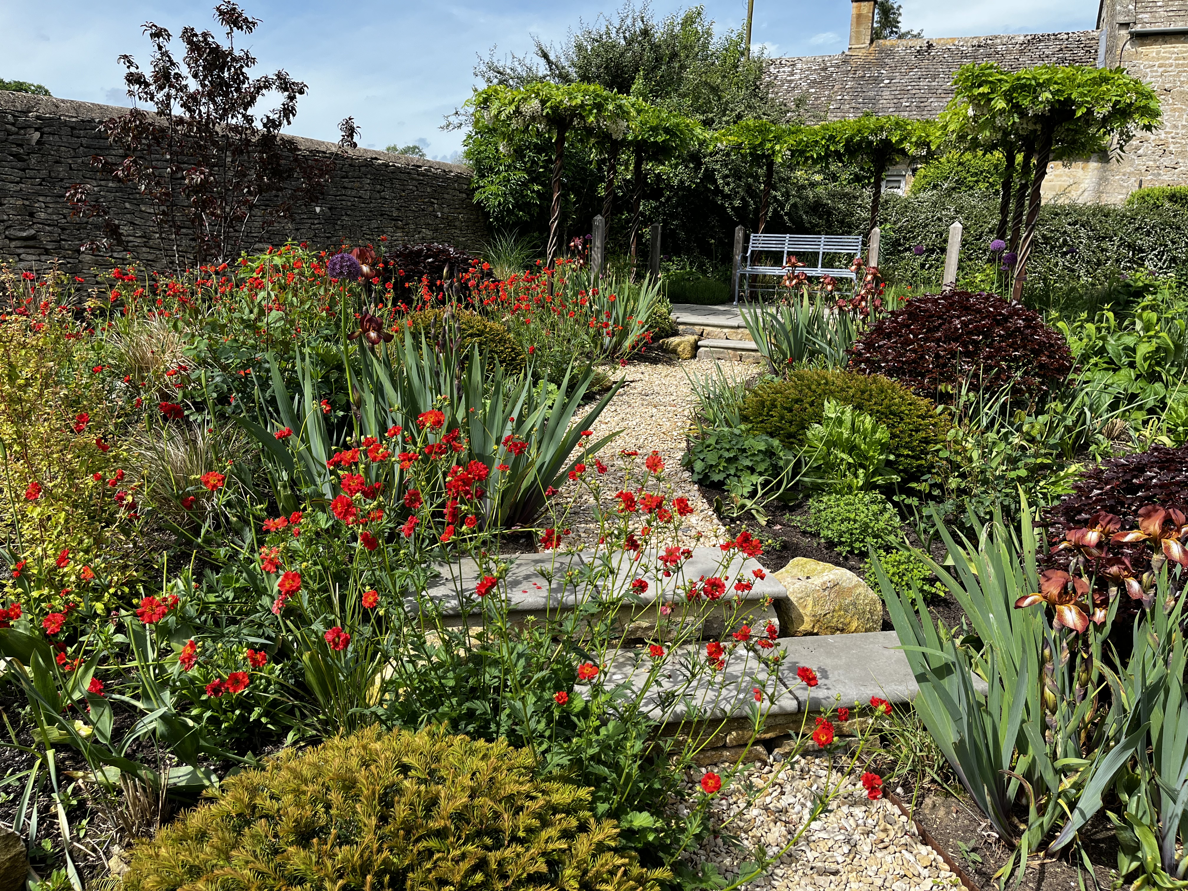 Sweeping red crocosmia and ornamental grass borders leading to a wisteria-clad pergola against a Cotswold stone wall